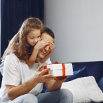 Na imagem, um homem sentado sobre um sofá com um presente nas mãos. Atrás dele tem uma garotinha dando um abraço pelas costas e tampando os olhos dele com as mãos para reforçar a surpresa.